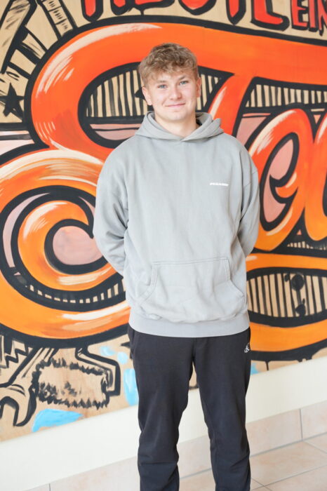 Das Bild zeigt einen jungen Mann vor einer bunten Wand stehend. Es handelt sich dabei um den Auszubildenden Mauerer Josef