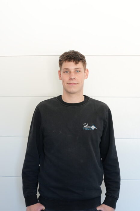 Das Bild zeigt einen jungen Mann vor einer weißen Wand stehend. Es handelt sich dabei um den Auszubildenden Fliesenleger Kevin