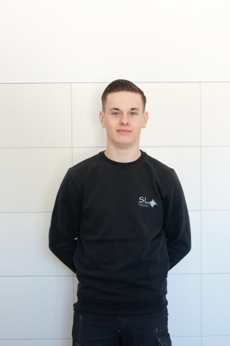 Das Bild zeigt einen jungen Mann vor einer weißen Wand stehend. Es handelt sich dabei um den Auszubildenden Fliesenleger Leon